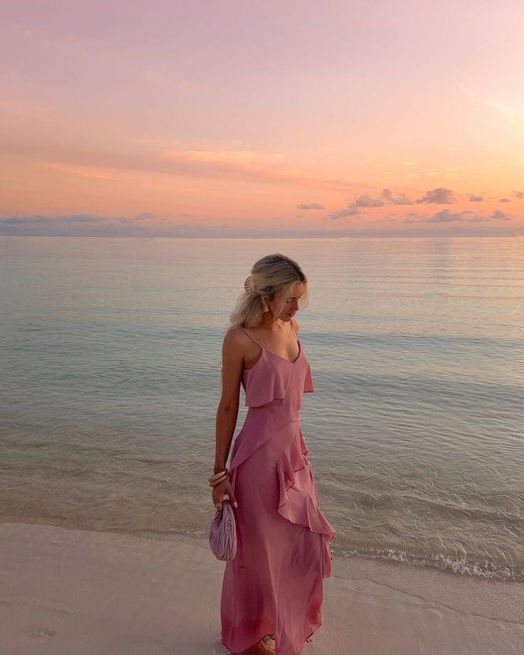 Woman in a pink dress stands on a beach at sunset, with a serene ocean backdrop and pastel sky.