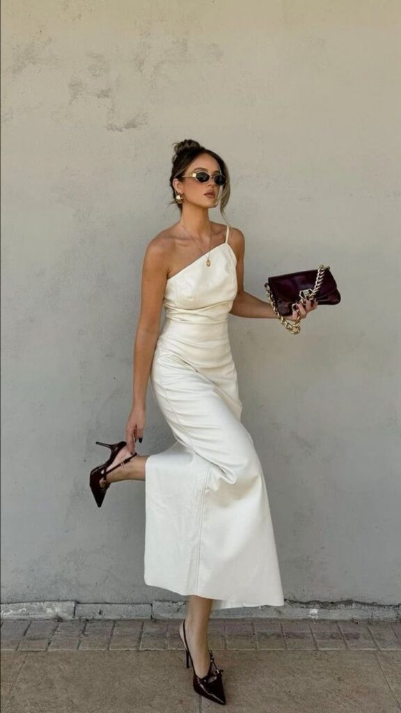 Woman in elegant white dress and heels posing with a chic purse against a neutral wall. Stylish fashion portrait.