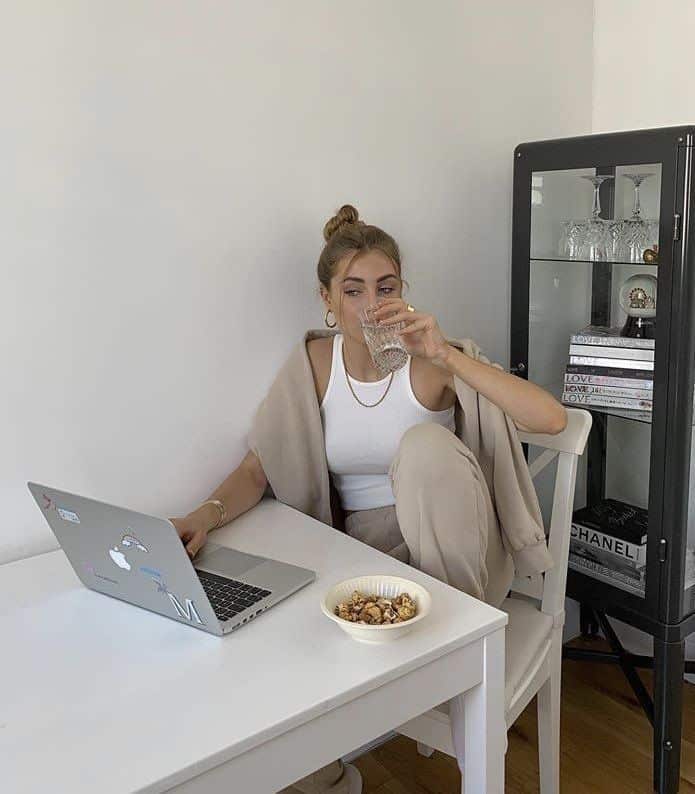 Woman working on a laptop at home, drinking water, with a bowl of cereal on the table for a healthy snack break.