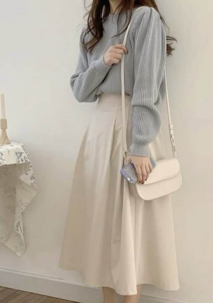 Woman wearing a light sweater and beige skirt, holding a cream bag in a minimalistic room.