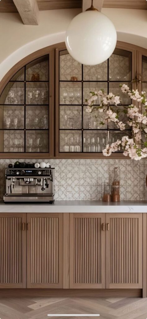 Modern kitchen with wood cabinets, glassware display, espresso machine, and floral arrangement.
