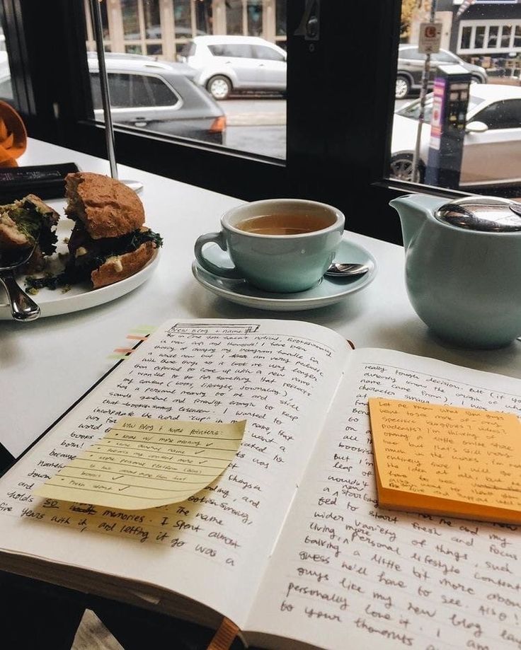 Open notebook with handwritten notes, accompanied by tea and a sandwich on a cafe table by the window.