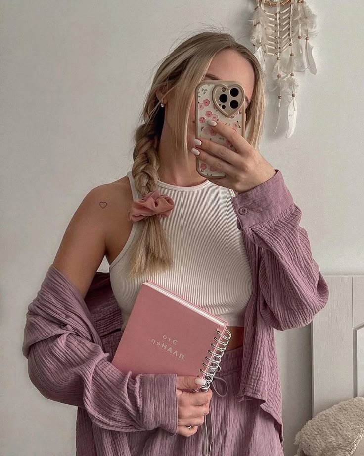 Woman in pink outfit holding journal, taking mirror selfie, with dreamcatcher hanging on wall in background.