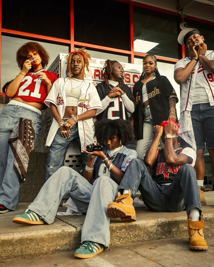 Group of stylish friends in sports jerseys and jeans pose outside a building, some seated, holding a camera.