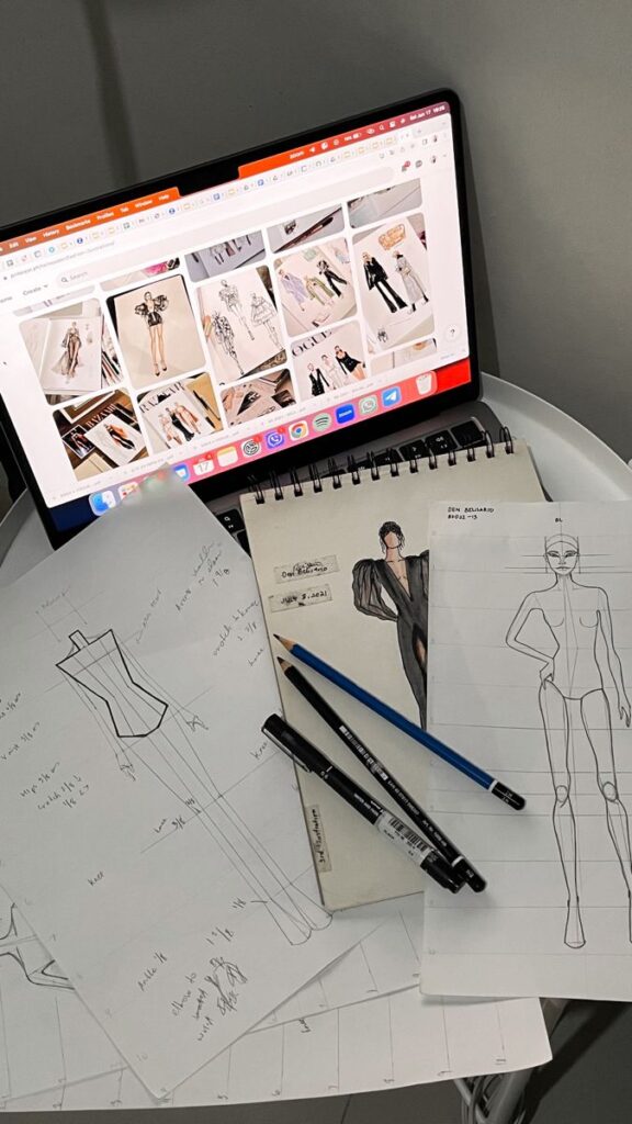 Fashion design sketches and laptop displaying fashion illustrations. Writing tools scattered on a white surface.