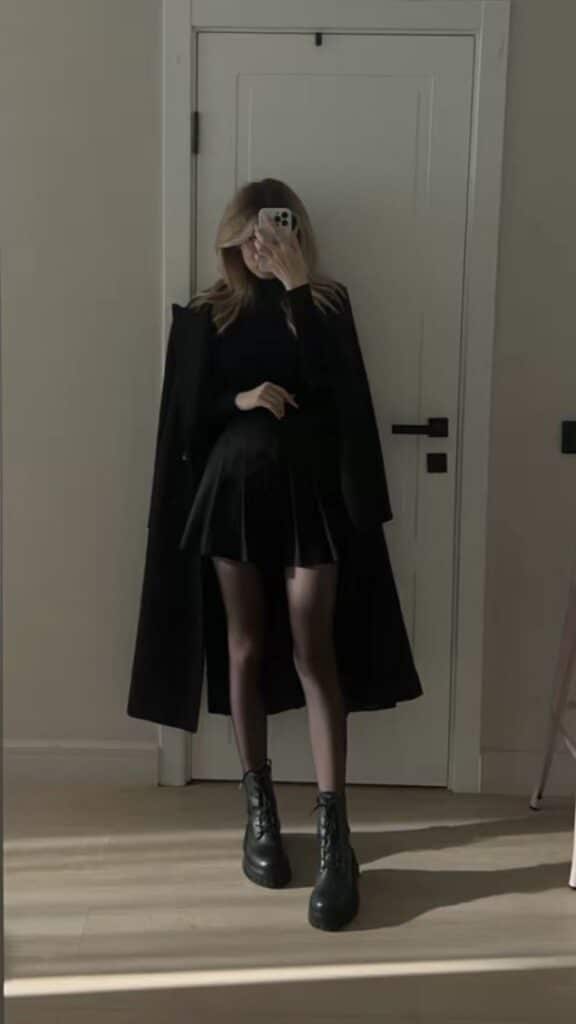 Stylish woman in a black outfit and boots taking a mirror selfie. Fashionable look with a pleated skirt and coat.