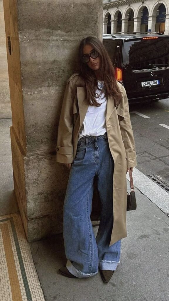 Woman in trench coat and wide-leg jeans leans against a city wall, showcasing casual street style fashion.