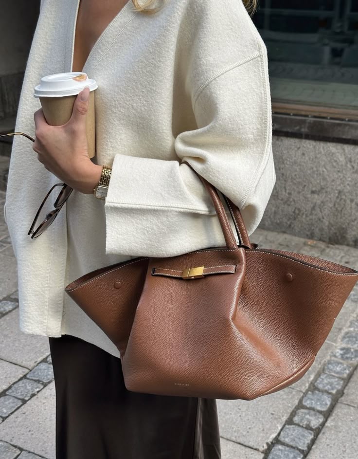 Woman holding coffee cup and brown leather handbag, wearing white coat and sunglasses; stylish street fashion.