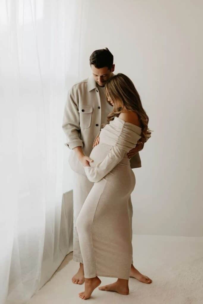 Expecting couple in soft neutral attire, sharing a loving moment next to a sunlit window.