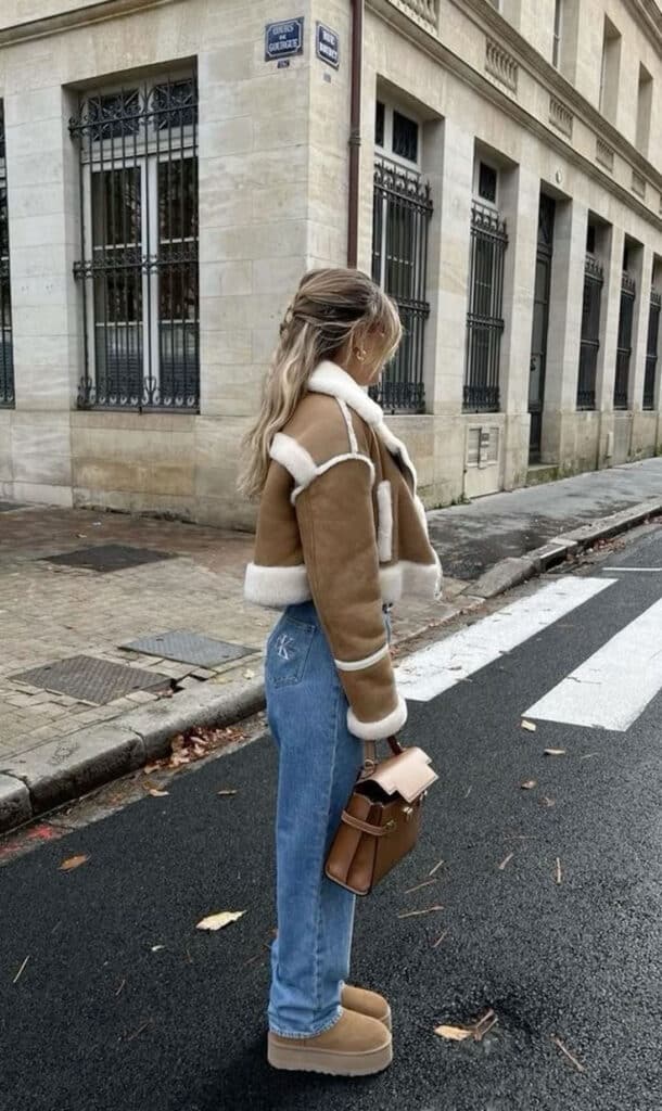 Fashionable person in a shearling jacket and jeans walks on a city street, holding a stylish leather handbag.