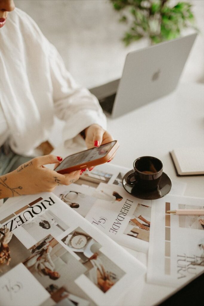 Person using phone at desk with fashion magazines, coffee, and a laptop. Modern workspace with greenery.