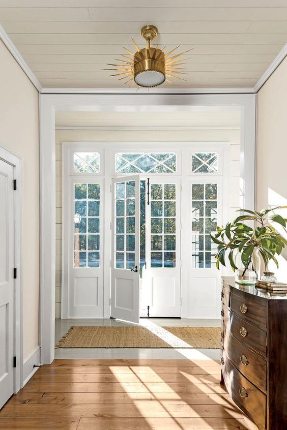 Bright entrance hall with glass doors and wooden floors, featuring a stylish chandelier and a wooden chest with a plant.