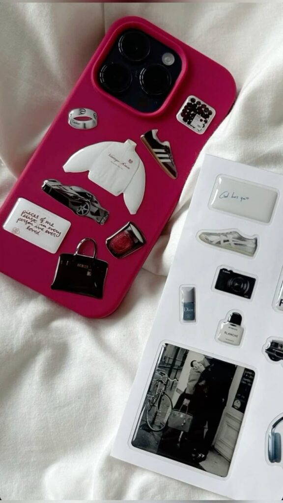 Pink phone case with trendy fashion stickers on a bed, next to a sheet of various stickers.
