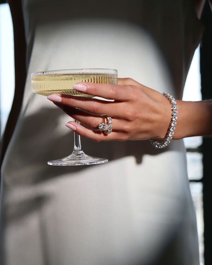 Elegant woman holding champagne coupe, showcasing diamond rings and bracelet, in a sophisticated setting.
