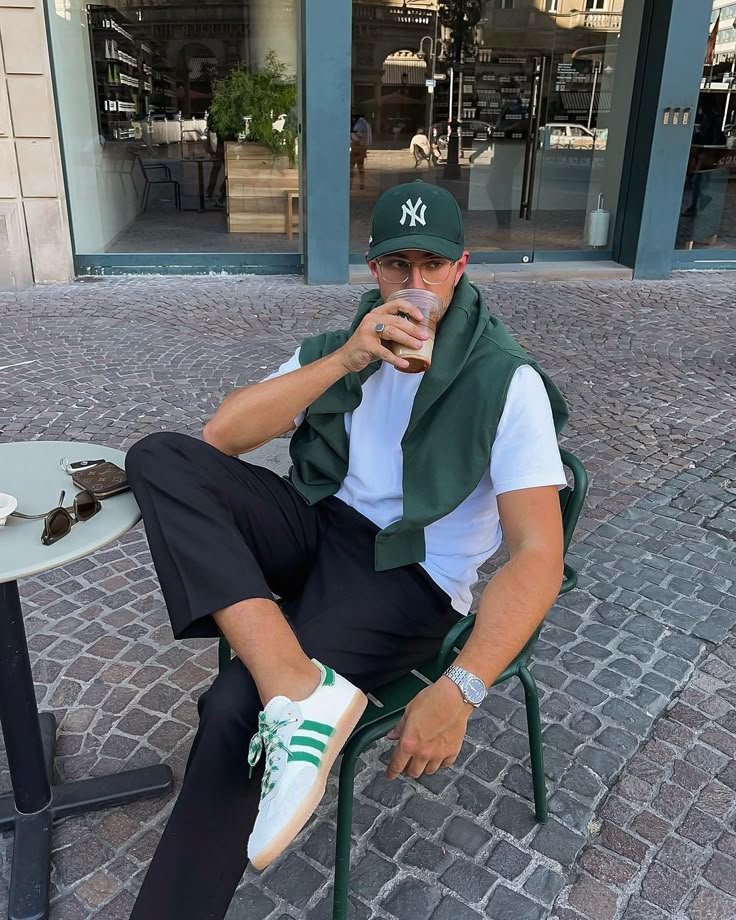 Man in casual outfit enjoying iced coffee outside a café, wearing a green cap, matching shoes, and a watch.