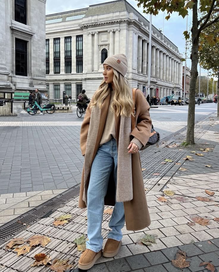 Woman in winter fashion, wearing a brown coat, jeans, and beanie, standing on a city sidewalk with historic buildings.