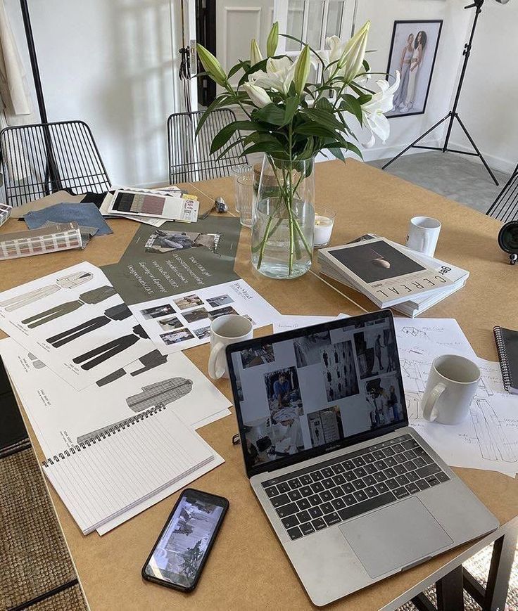 Fashion design workspace with laptop, sketches, flowers, and coffee cups on a table, ready for creative work.