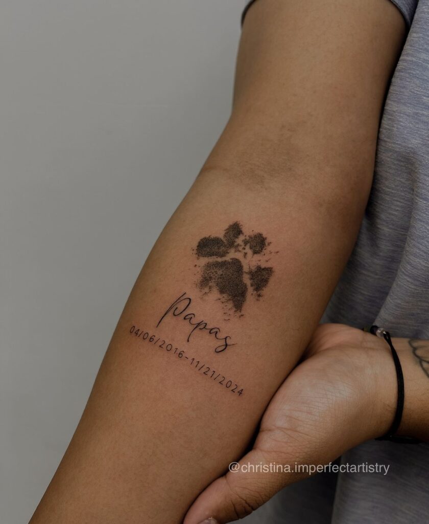 Arm tattoo of a paw print with Papas and dates, symbolizing a pet memorial on a person's forearm.