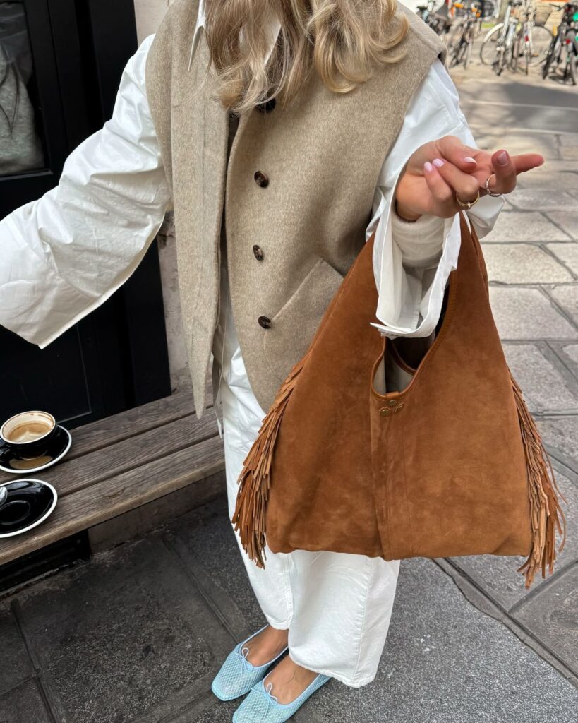 Woman in stylish beige vest holds brown fringe bag, standing by coffee cups on a wooden bench.
