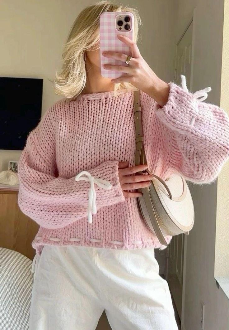 Woman in pink knit sweater and white pants takes a mirror selfie with a plaid phone case, carrying a beige round bag.