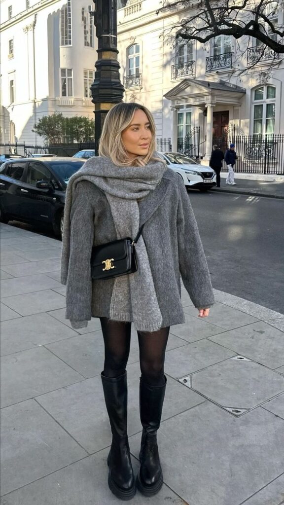 Woman in stylish winter outfit with gray coat and scarf, black boots, and purse, standing on city street.