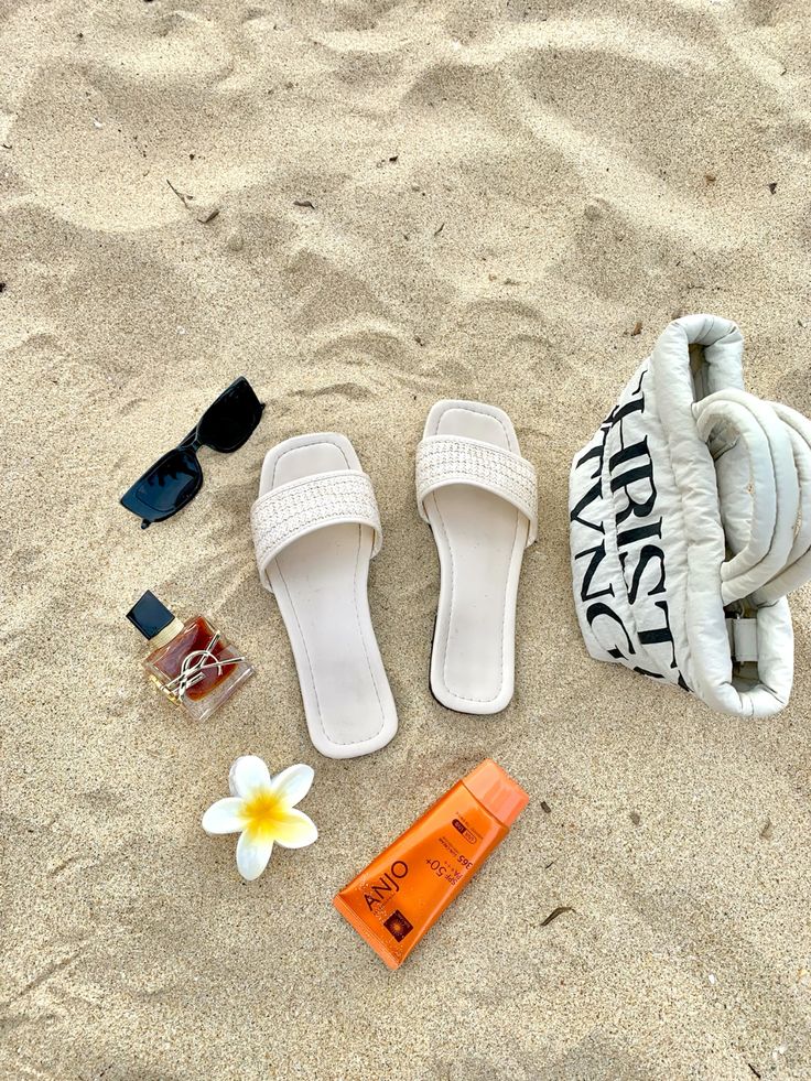 Beach essentials on sand: slides, shades, fragrance, plumeria flower, sunscreen, and a stylish tote bag.