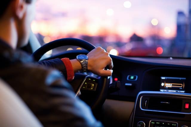 Person driving a car at sunset, with focus on the steering wheel and dashboard, city lights in the background.