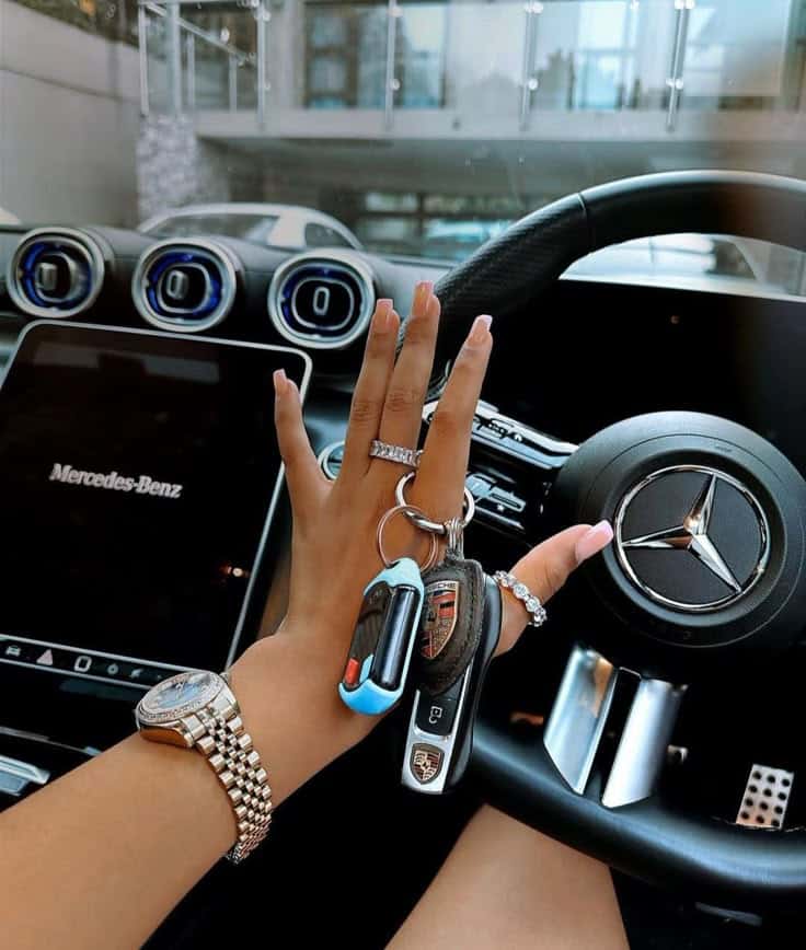 Hand with luxury watch and rings holding Porsche keys inside a Mercedes-Benz car.
