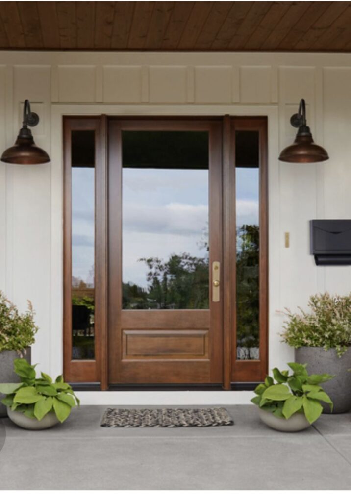 f709143e-a791-4739-91ac-392010e0b5cf Front porch with wooden door, potted plants, doormat, and wall lights. | Sky Rye Design Front porch with wooden door, potted plants, doormat, and wall lights.