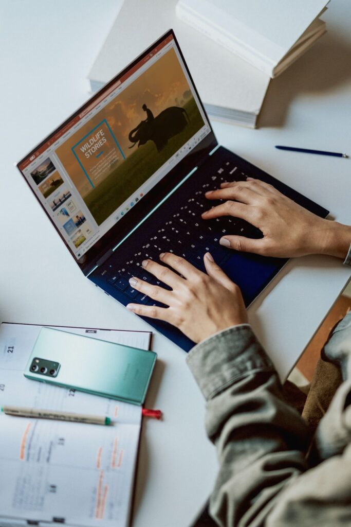 Person typing on a laptop with a wildlife presentation next to a phone and planner on a desk.