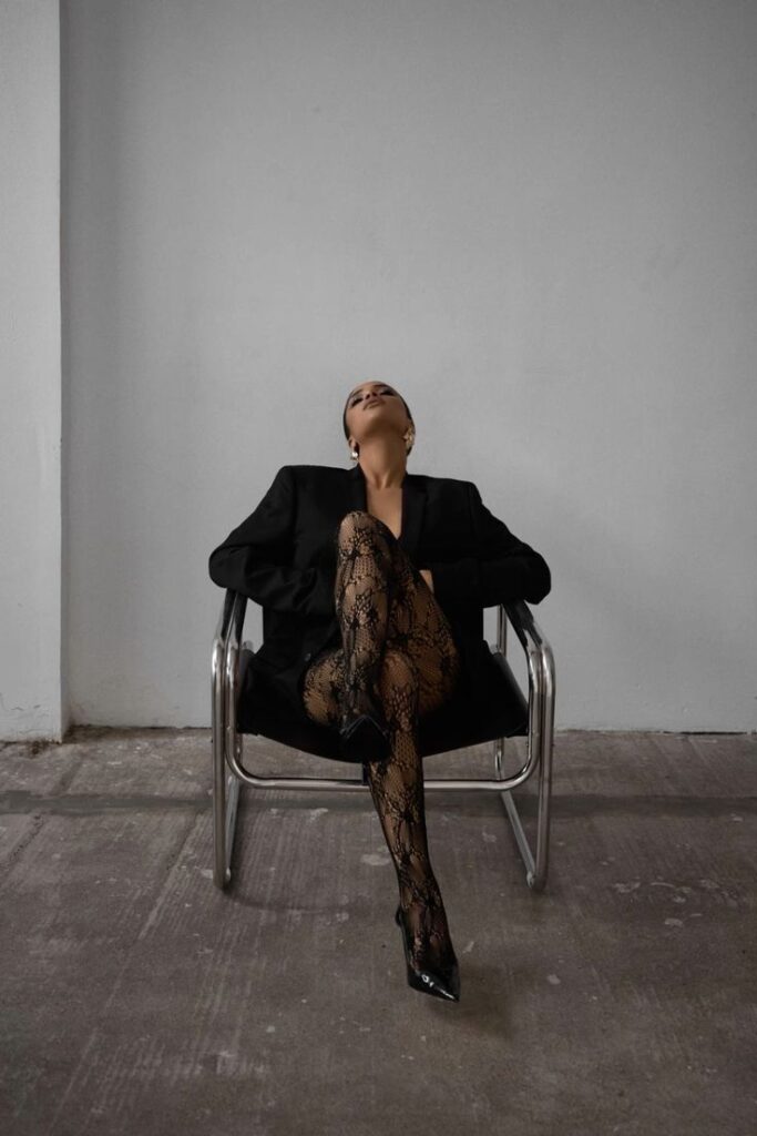 Fashionable woman in lace tights and blazer, seated on a modern chair in a minimalist setting.