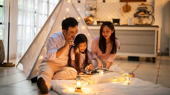 Family reading book together in cozy living room, sitting by a teepee tent with string lights and smiles.