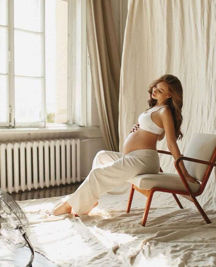 Pregnant woman in white outfit sitting on chair by sunlit window, serene indoor setting.