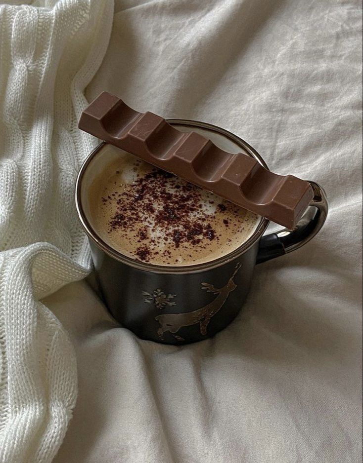 Cozy hot chocolate with cocoa sprinkle and chocolate bar on a black mug, set on soft white fabric.