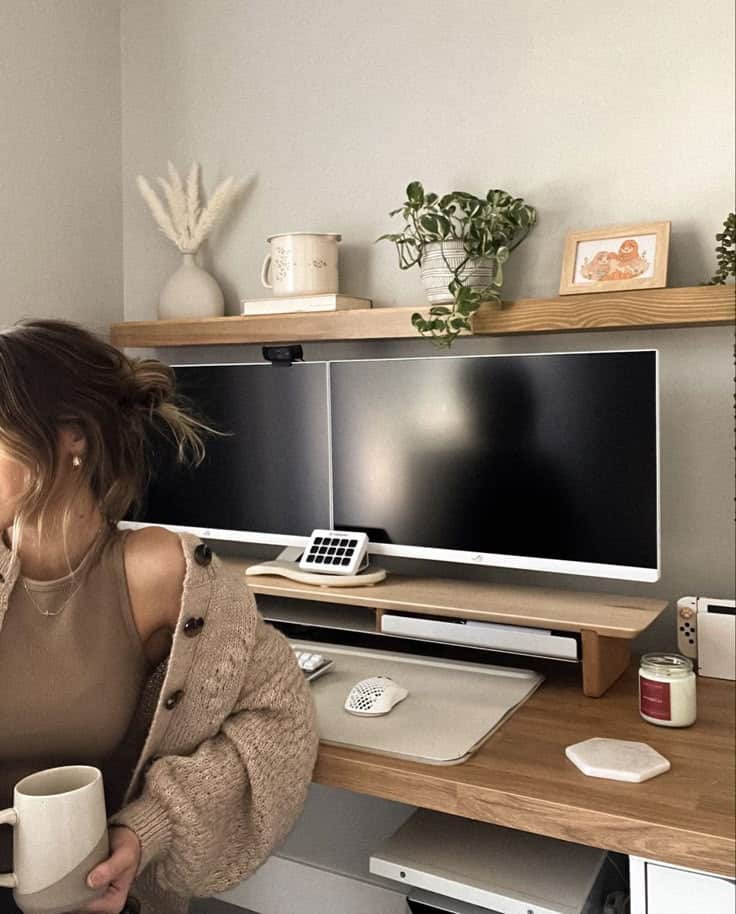 Cozy home office with dual monitors, plants, and a person holding a mug near a wooden desk setup.