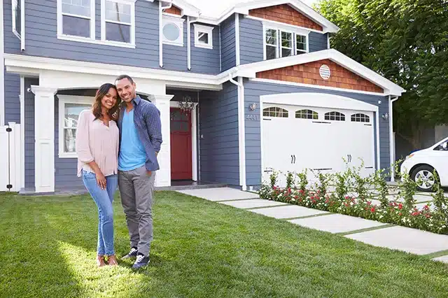 Couple smiling in front of a modern blue house with a well-maintained lawn and garden.