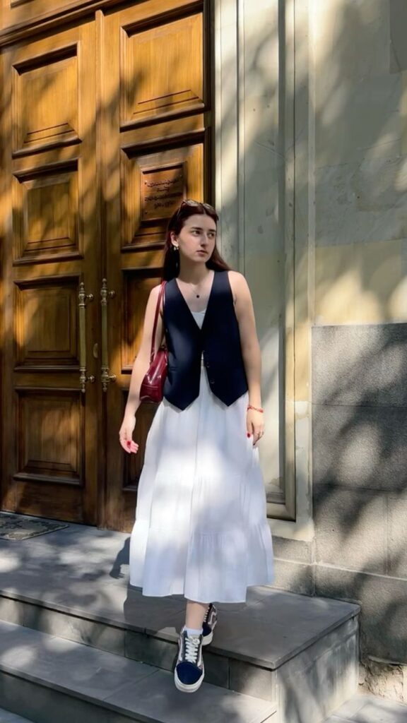 Woman in a white dress and black vest stands in sunlight by a wooden door, holding a red bag.