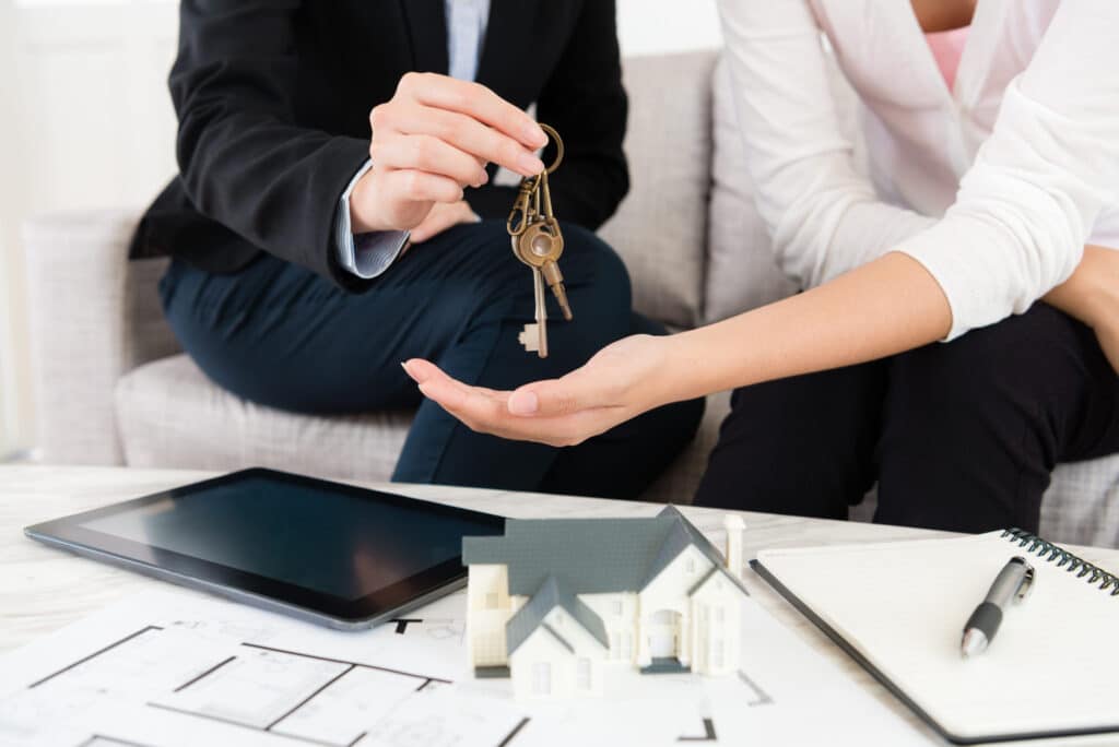 Real estate agent handing over house keys, with tablet, model home, and floor plans in view.