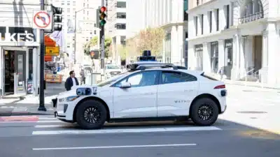 Autonomous white Waymo vehicle at a city intersection, stopped at a pedestrian crossing during the day.
