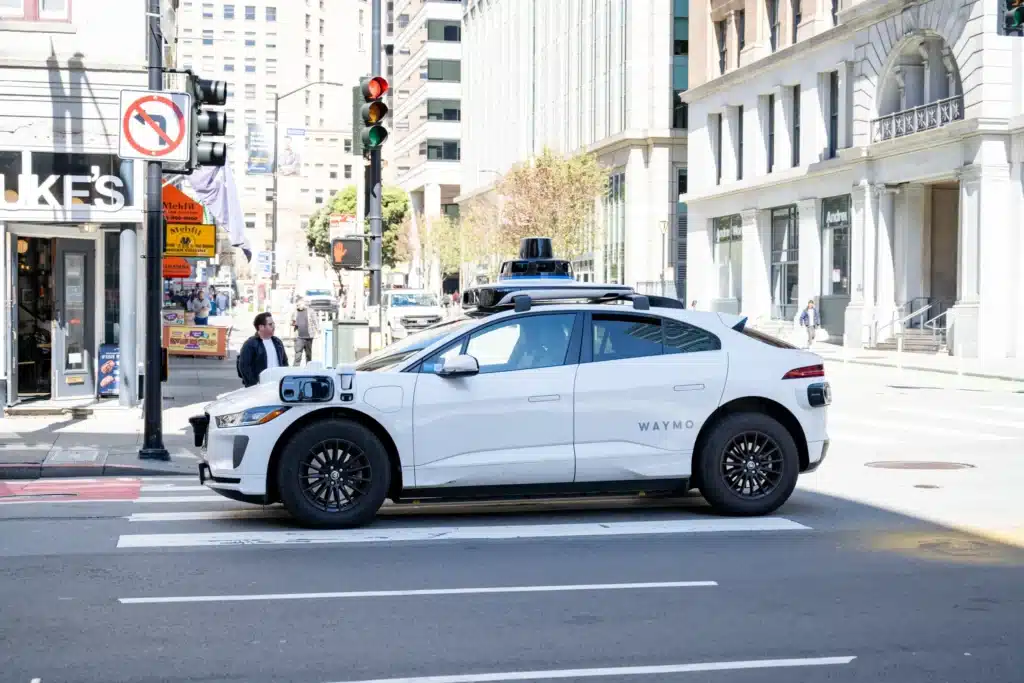 Self-driving Waymo car stopped at a city intersection near buildings and a traffic light.