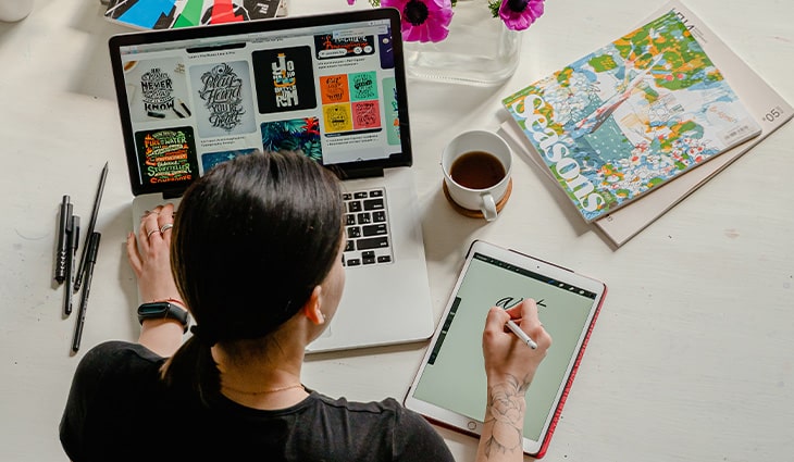 Graphic designer working on laptop and tablet, creating digital art. Coffee cup, magazine, and pens on white desk.