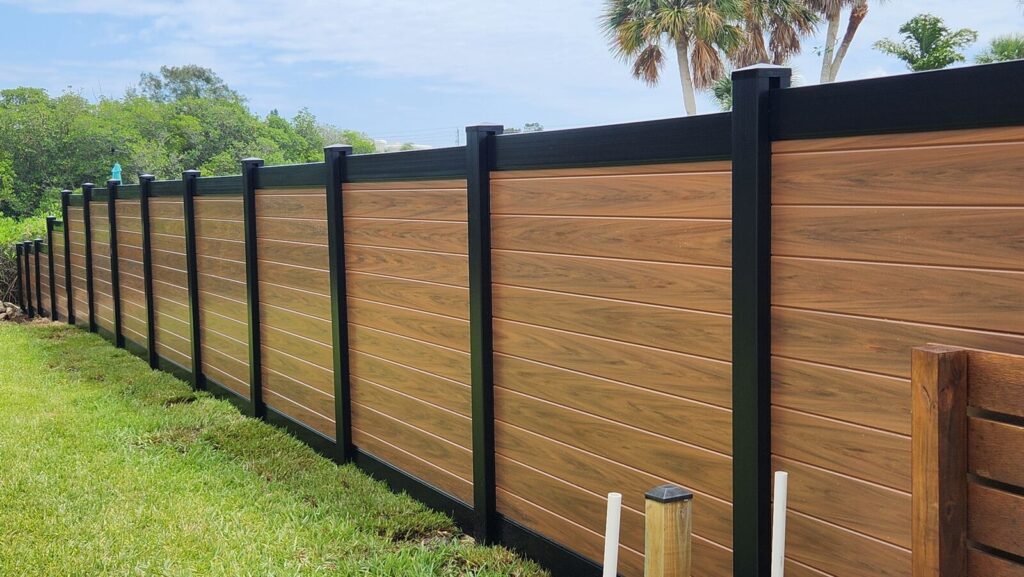 Stylish brown composite fence with black pillars in a lush green garden setting under a clear blue sky.