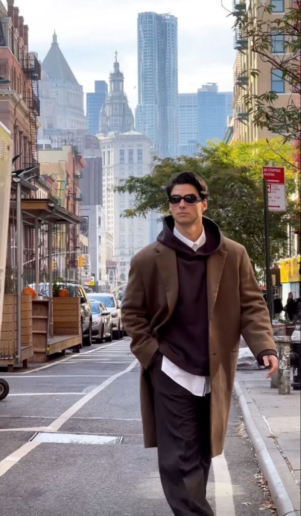 Man in brown coat walks confidently in urban street with skyscrapers in the background. Stylish city fashion.