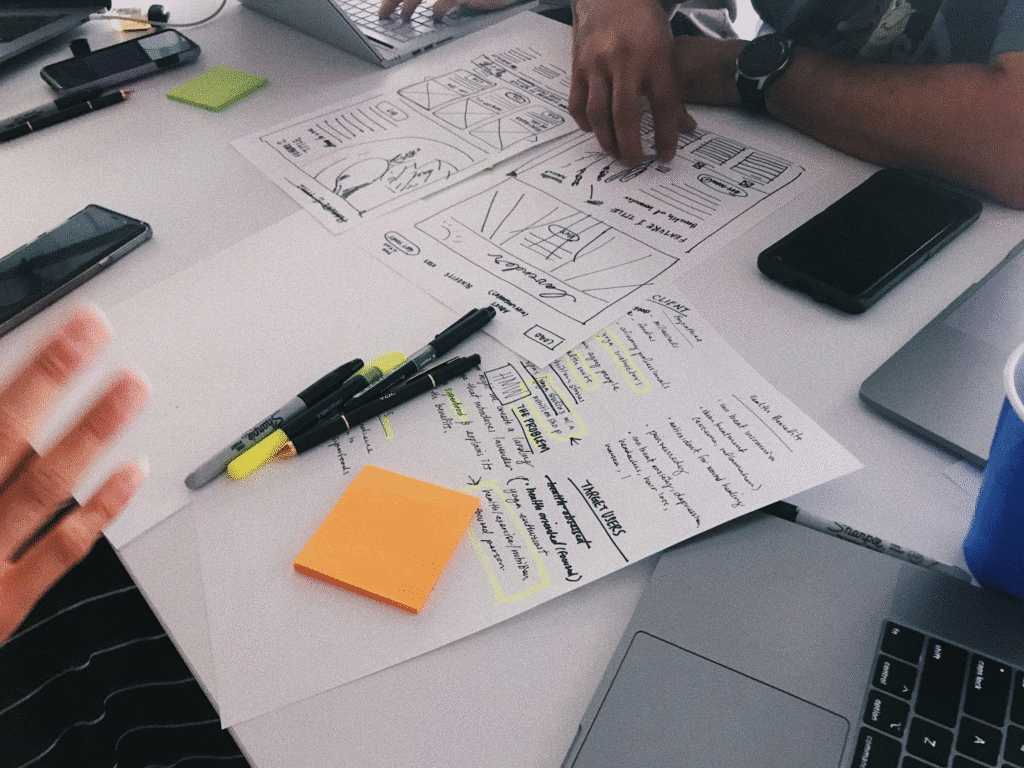 image Team collaborating on design plans and notes with laptops and phones on a desk. | Sky Rye Design Team collaborating on design plans and notes with laptops and phones on a desk.