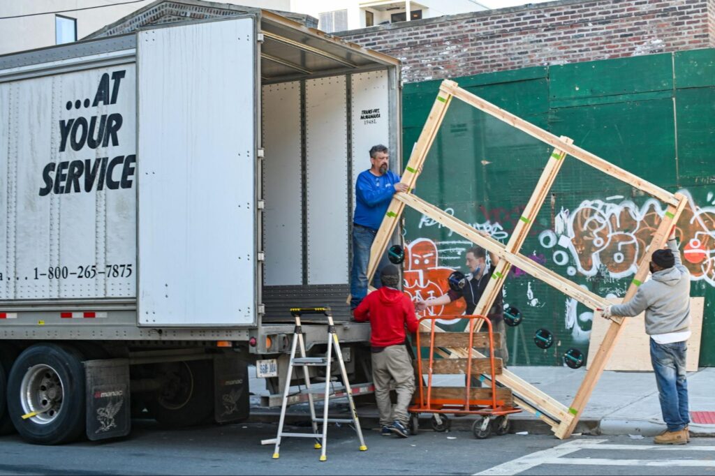 Workers loading large wooden frame onto truck near graffiti-covered wall.