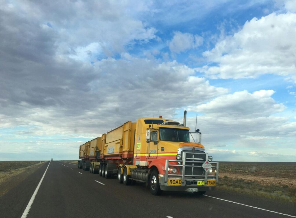 Yellow road train truck on a flat desert highway under a partly cloudy sky.