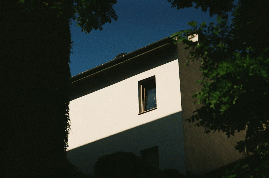 White building facade with open window, surrounded by trees, casting dramatic shadows under a clear blue sky.