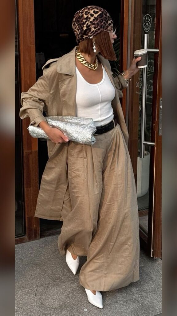 Stylish woman in beige trench coat, leopard print headscarf, holding coffee and silver clutch outside a café.