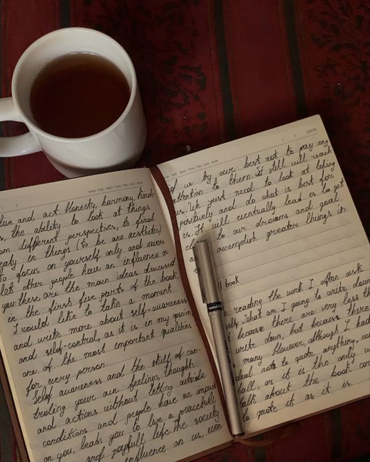 Open notebook with handwritten text and pen, accompanied by a cup of tea on a dark wooden table.