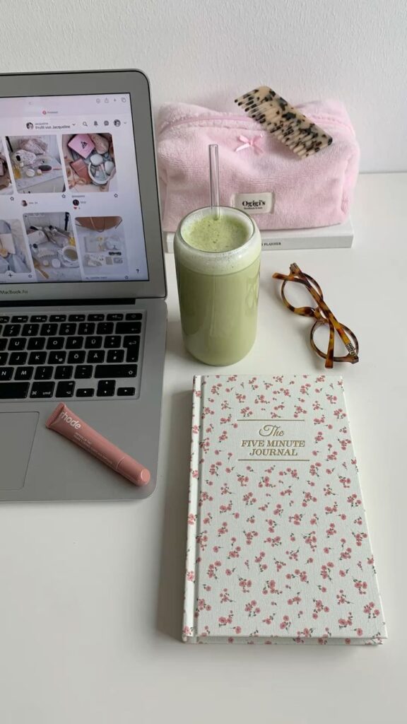 Laptop, green smoothie, floral journal, glasses, and pink makeup on a desk. Cozy and productive workspace setup.